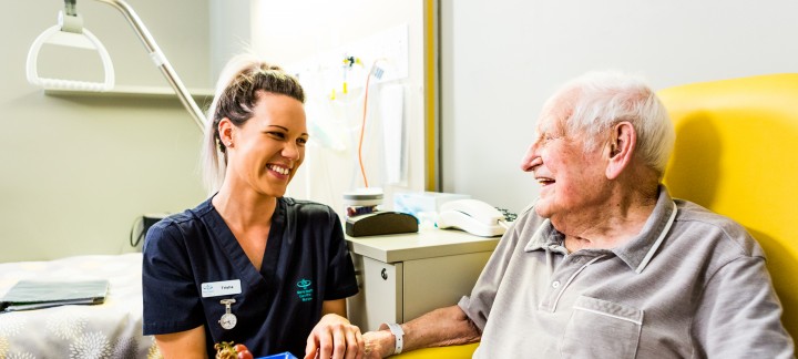 An elderly man sits in a yellow chair at a table with a bowl of fruit, holding the hand of a young female Mercy Health worker. The man and woman are smiling at each other.