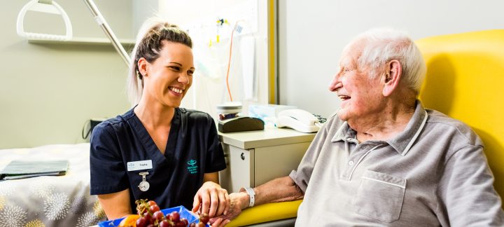 An elderly man sits in a yellow chair at a table with a bowl of fruit, holding the hand of a young female Mercy Health worker. The man and woman are smiling at each other.