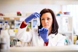 A medium close up of a woman in a lab wearing blue gloves and white lab coat holding a test tube.