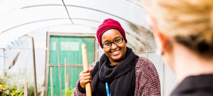 An over the shoulder photo of a female Mercy Health worker in a green house holding a gardening tool and smiling at a colleague